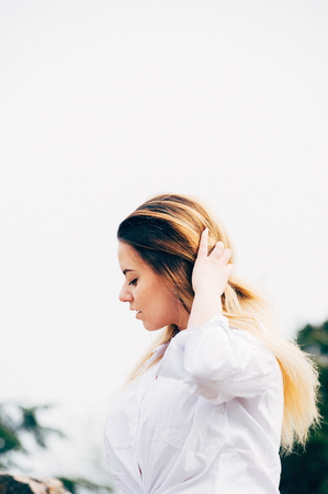 A portrait of a pretty young long haired girl in white shirt, blurred background with green trees. Half face view, eyes looking down, touching hair with handの写真素材