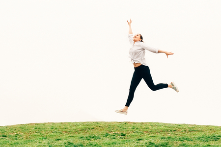 A pretty young long haired woman in white shirt and black jeans, jumping above green grass against sky backgroung, freedom conceptの写真素材