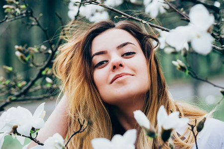 A portrait of a pretty young long haired girl in a garden enjoying blooming magnolia trees with white flowers, romantic look, closeup view, blurred background and foreground with branchesの写真素材