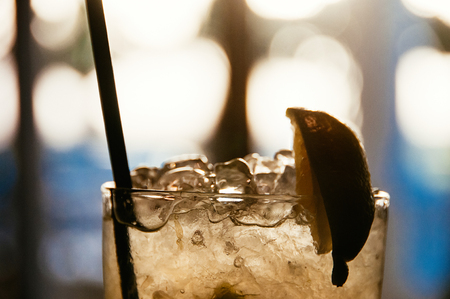 a closeup view of an old fashion glass with cocktail, ice cubes, black sipping straw and lime, blurred bokeh in the backgroundの写真素材