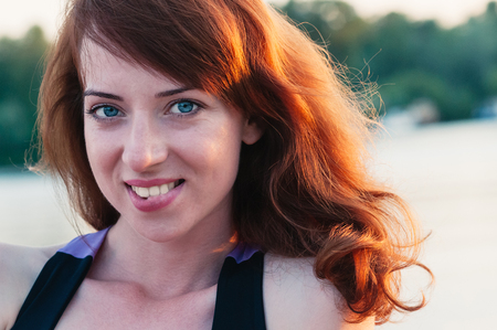 Close up portrait of a young beautiful ginger haired woman, biting her lip in a smile, white teeth, looking at camera, on water summer nature backgroundの写真素材