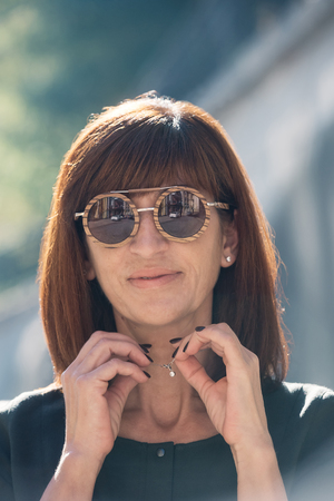 Street style Fashion portrait pretty middle age woman in stylish dark luxury garment, smiling, sun glasses with wooden frame. Sunny day, city lifestyle. Motion blurの写真素材
