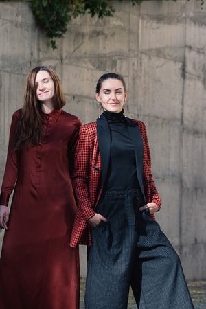 Street style Fashion portrait two charming girls posing. Brown dress, Black and tile red jacket. Summer Spring Fall collection beautiful look. Togetherness, friendship, party conceptの写真素材
