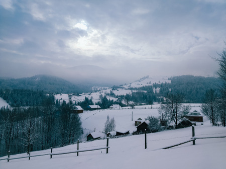 Winter countryside mountain landscape with wooden houses covered with snow, forests in the misty distant background. Picturesque and peaceful wintry scene. European resort location. Blue toningの写真素材