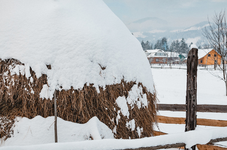 Big dry haystack covered with snow cap. Winter rural landscape, village or farm, snow-capped mountains and forest in the backgroundの写真素材