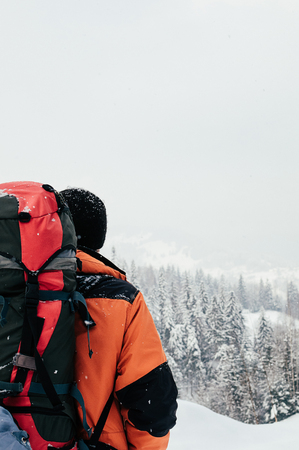 Rear view of tourist male enjoying beautiful mountain forest landscape on snowy winter day. Orange garment, red backpack. Hiking travel extreme lifestyle conceptの写真素材