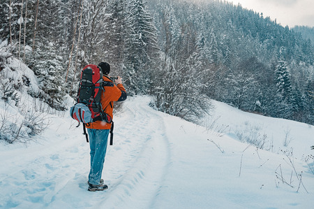 Rear view of tourist taking photo of mountain forest landscape on snowy winter day. Blue jeans, orange garment, red backpack. Hiking travel extreme conceptの写真素材