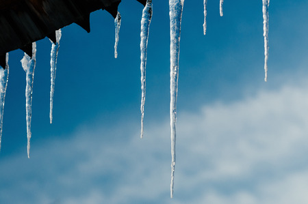 Background of long bright transparent icicles in the sunlight, hanging from wooden roof. Spring blue sky. Selective focus. Copy space for text and designの写真素材