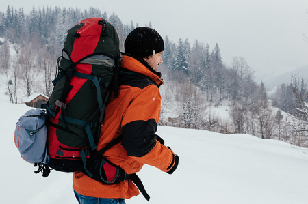 Tourist man walking through snow on winter day, smiling, mountain forest landscape. Blue jeans, orange garment, red backpack. Hiking travel extreme concept. Selective focusの写真素材