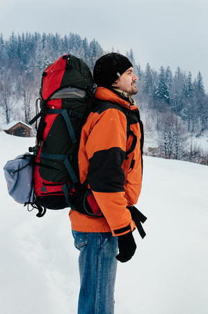 Tourist man breathing, enjoying fresh air and view of mountain forest landscape on a snowy winter day. Blue jeans, orange garment, red backpack. Hiking travel extreme conceptの写真素材