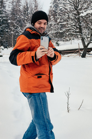 Young man taking picture with his smartphone, smiling. Blue jeans, orange garment .Winter day, snow hill, trees, countryside. Fresh air. Fun and leisure conceptの写真素材