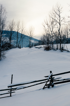 Wooden fence covered with snow cap. Winter rural landscape, village or farm, snow-capped mountains and forest in the sunset background. Vertical imageの写真素材