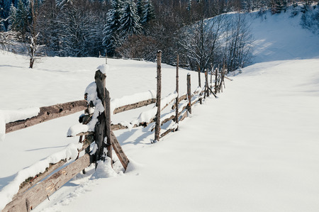Wooden fence covered with snow cap. Winter rural landscape, village or farm, sunny day, blue shadows, mountains and forest in backdropの写真素材