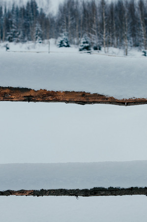 Wooden fence beams covered with snow cap. Winter rural landscape, village or farm, cloudy day, forest in backdrop on hill. Vertical imageの写真素材