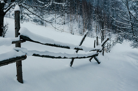 Wooden fence beams covered with snow cap. Winter rural landscape, village or farm, cloudy day, forest in backdrop on hillの写真素材