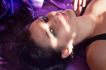 Beauty portrait of brunette woman model lying on purple silk texture as backdrop, natural make-up. Fashion shiny highlighter on skin, sexy gloss lips. Glamour, sensual, seductive look, perfect skinの写真素材