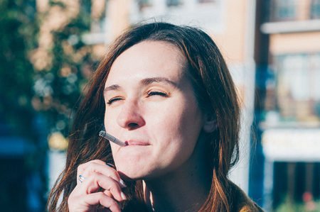 Real young woman smoking a cigarette in the street, inhaling, squinting her eyes into the sun.  Selective focus. Addiction, bad habits, everyday life conceptの写真素材