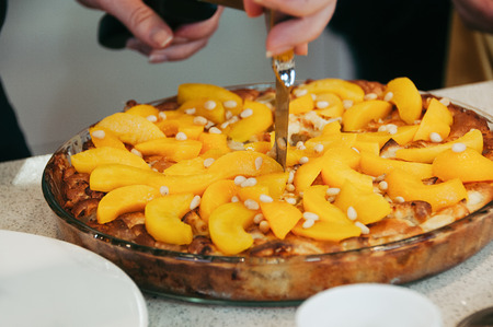 Delicious vegan peach cake decorated with pine nuts, whole, in round glass pan, on table. Selective focus. Colorful healthy juicy. Side view, female hand with knife cutting the cakeの写真素材