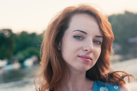 Close up portrait of a young beautiful ginger haired woman, looking at camera, blue eyes and clothes, sunset light, summer nature background, motion blur, messy hair by the wind. Street fashionの写真素材