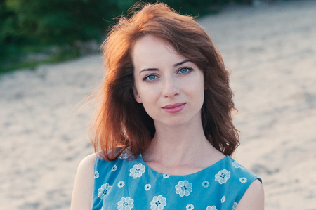 Close up portrait of a young beautiful ginger haired woman, looking at camera, blue eyes and clothes, sunset light, summer nature background, motion blur, messy hair by the wind. Street fashionの写真素材