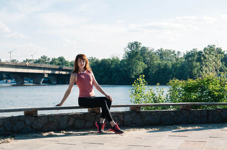 Beautiful fit young woman sitting on a bench, wearing tight training clothes, smiling, long hair, river background. Healthy lifestyle in the city, keep fit, enjoy life conceptの写真素材