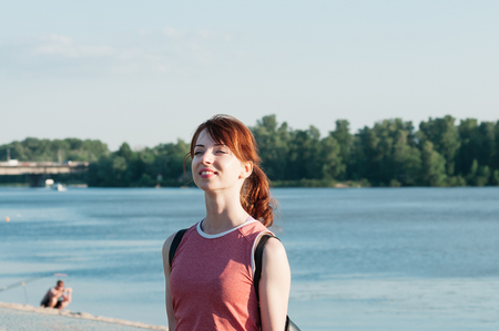 Close up portrait of a young beautiful ginger haired woman, perfect smile and white teeth, red top, looking into distance, sunny, water summer nature background, copy space. Travel conceptの写真素材