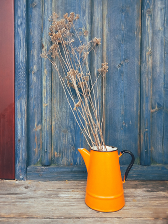 Dry flowers on long stems in an orange metal jug, on blue wooden veranda background. Daylight. Still life in rustic style. Countryside lifestyle concept. Copy spaceの写真素材