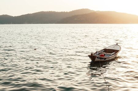 Traditional fishing boat docked in the sea at sunset. Old wooden row boat in the sea, mount island in the background in the sun rays at sunset. Travel, vacation, getaway, loneliness, solitude conceptの写真素材