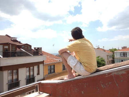 a young man sitting on the roof of a house enjoying the view of the old city, pointing at seagulls flying in the cloudy sky on a summer dayの写真素材