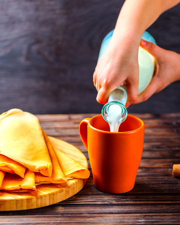 Children's hands pouring milk from a glass bottle. Pancakes on a wooden Board nearby. Wooden dark background. Concept of homemade Breakfastの写真素材