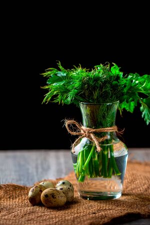 Bunch of parsley and dill in a glass vase. Quail eggs near on a dark background. Concept of spring. Vertical imageの写真素材