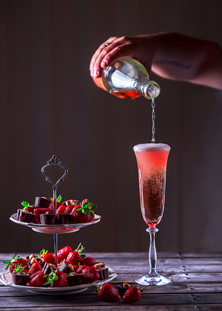 Sparkling pink wine is poured in glass. Stand with strawberries and sweets on a wooden table.  Delicious dessert. Atmosphere of luxury and a romantic evening. Selective focusの写真素材