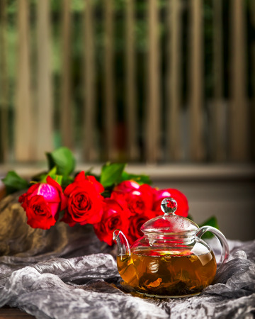 Glass teapot. A teapot with tea on the table. Glass teapot on a dark background. A bouquet of roses in the background. A bouquet of red roses next to a glass kettleの写真素材