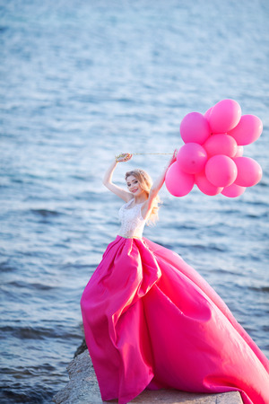 young beautiful girl in a pink dress holding pink balloons on the beachの写真素材