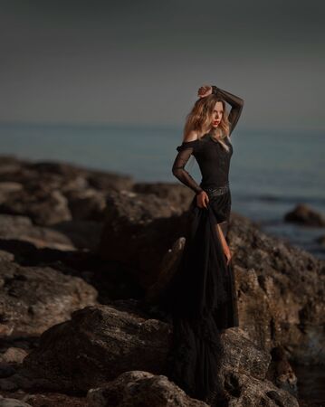 Young woman in long transparent black dress among beach and sea. Wind blows hem of her dress.の写真素材