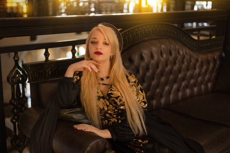 Portrait of a beautiful blonde girl with long hair in a black dress sitting on a leather sofa.