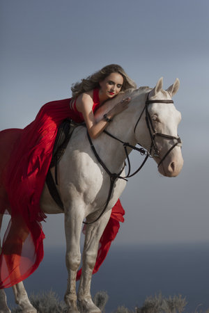 Cheerful bright girl leans on the horse, eyes closed, hand on the hair. Fashion blonde in the red dress. Travel around the countryside.の写真素材