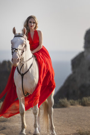 Cheerful bright girl leans on the horse, eyes closed, hand on the hair. Fashion blonde in the red dress. Travel around the countryside.の写真素材