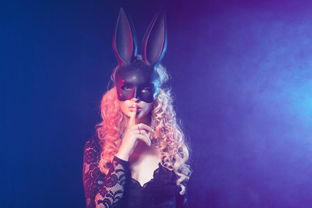 Young woman in black rabbit or hare fancy mask and black dress. Blonde female with long hair and hand near face. Female on smoke and metal wall background.の写真素材
