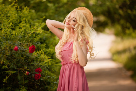 Close up portrait of an attractive young woman in summer dress and straw hat holding carnations bouquet and looking over her shoulder isolated over pink backgroundの写真素材