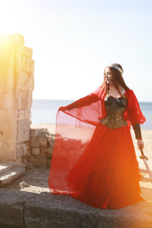 Beautiful red haired girl in metal medieval armor dress with sword standing in warlike pose and looking at camera with fearless. Fairy tale story about warrior. Warm art work.の写真素材