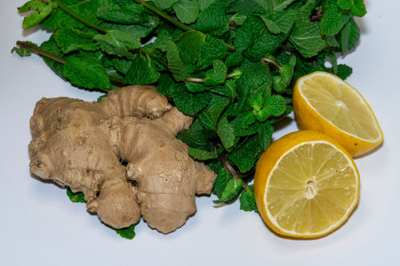 A close-up of a composition of raw ginger root, fresh green mint, and two bright lemon halves,...