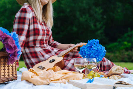 Girl in red checkered dress and hat sitting on white knit picnic blanket plays ukulele and drinking wine. Summer picnic on sunny day with bread, fruit, bouquet hydrangea flowers. Selective focusの写真素材