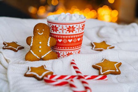 Christmas postcard with gingerbread man red mug with christmas ornament on light bokeh from Xmas tree and red striped holidays candies. Xmas candies and cup with marshmallow on white knit backgroundの写真素材