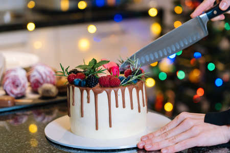 Person slicing homemade christmas fruit cake. Happy New Year and Merry Christmas Background. Winter decorations over dark background with defocus xmas tree light. Selective Focus.の写真素材