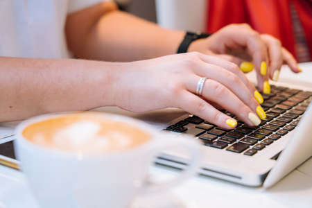 Close up cup of cappuccino coffee and hands woman sitting in office coworking and typing on her laptop keyboard computer. Desk with a phone, notebook, glasses and a cup of coffee. Business concept.の写真素材