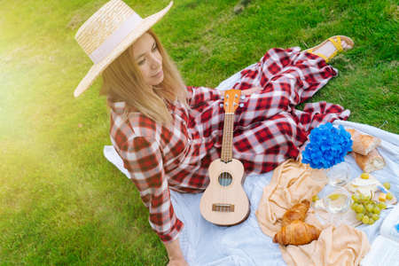 Girl in red checkered dress and hat sitting on white knit picnic blanket plays ukulele and drinking wine. Summer picnic on sunny day with bread, fruit, bouquet hydrangea flowers.の写真素材