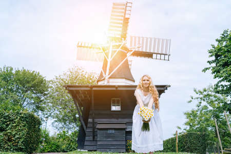 Woman dressed on traditional Dutch dress, wooden shoes yellow clogs klompen holding bouquet of Chamomile flowers with windmill on background. Retro vintage and countryside netherlands conceptの写真素材
