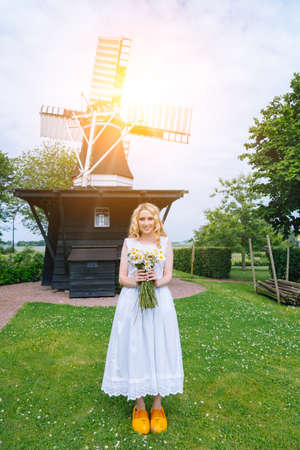 Woman dressed on traditional Dutch dress, wooden shoes yellow clogs klompen holding bouquet of Chamomile flowers with windmill on background. Retro vintage and countryside netherlands conceptの写真素材