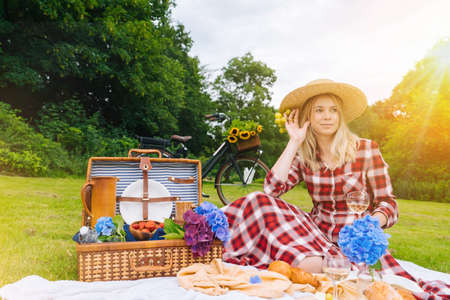 Girl in red checkered dress and hat sitting on white knit picnic blanket reading book and drinking wine. Summer picnic on sunny day with bread, fruit, bouquet hydrangea flowers. Selective focus.の写真素材